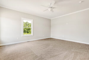 Bright, empty carpeted room with a window, ceiling fan, and light gray walls.