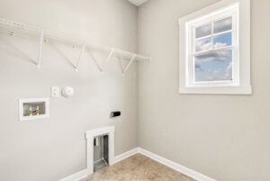 Laundry room with beige walls, empty wire shelf, washer hookups, and a window showing blue sky with clouds.