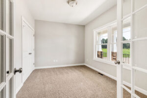 Bright, empty room with beige carpet, white walls, double doors, and a large window showing a green outdoor scene.