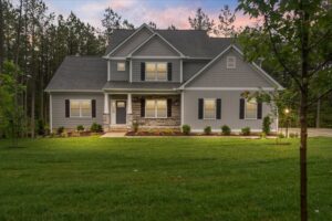 Two-story gray house at Shirley Commons with front lights on, large lawn, shrubs, and trees at sunset.