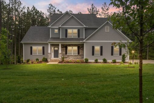 Two-story gray house at Shirley Commons with front lights on, large lawn, shrubs, and trees at sunset.