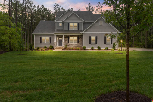 Two-story gray house at Shirley Commons with lights on, surrounded by grass and trees at dusk.
