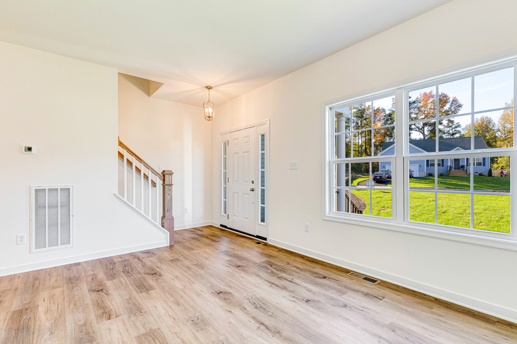 Bright, empty room in Poplar Village’s Lot 29 with wood floors, large window, staircase, and abundant natural light.