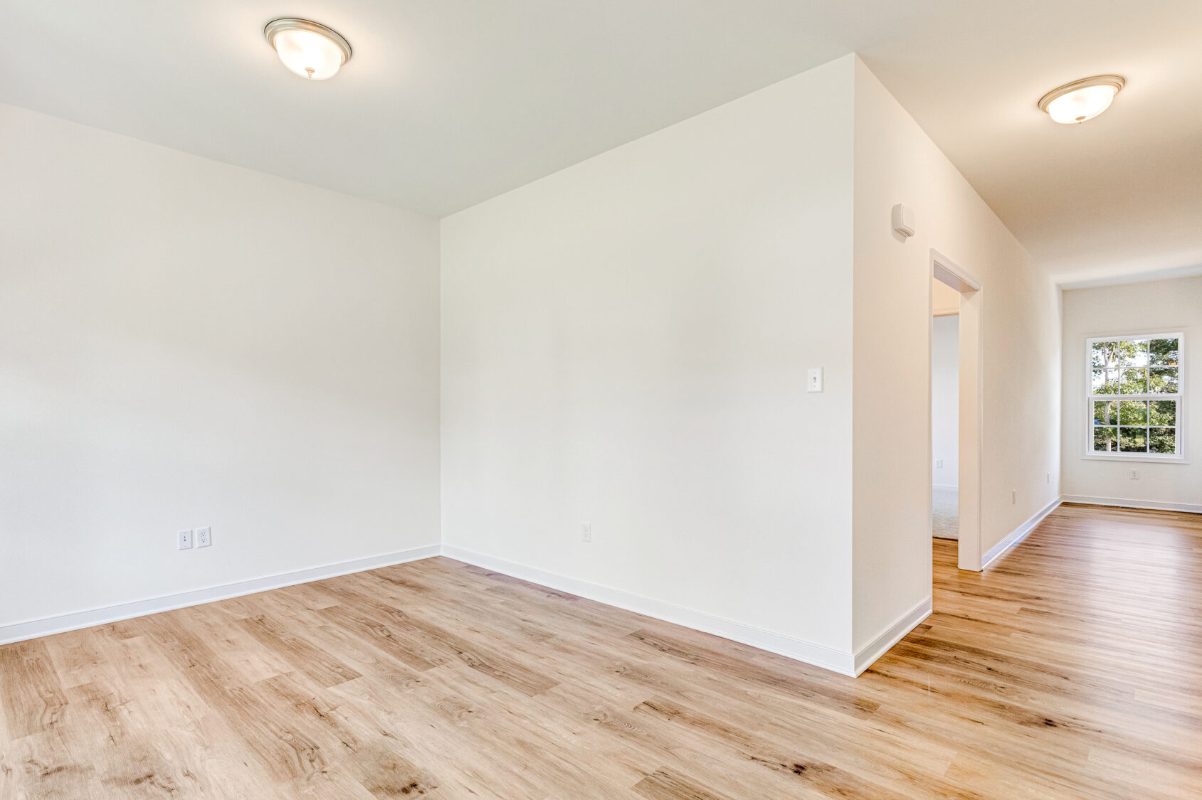Bright, empty room in Poplar Village with white walls, wood flooring, ceiling lights, and a distant window.