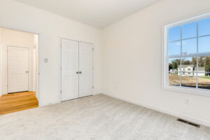 Bright, empty room with light carpet, white walls, double closet in Poplar Village Lot 29, window overlooks houses.