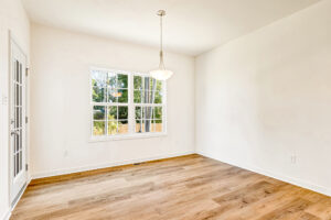 Bright empty room with wood flooring in Lot 29, Poplar Village; large window, glass door, and hanging light fixture.