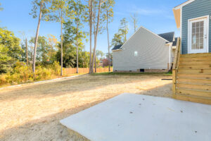 Backyard at Lot 29 in Poplar Village with bare ground, wooden stairs, concrete slab, and tall trees surrounding a neighbor.