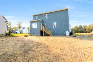 Blue two-story house on Lot 29 in Poplar Village with wooden stairs leading to a backyard with dry grass and distant trees.