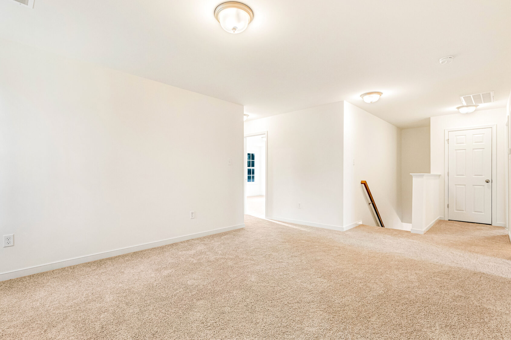 Bright, empty carpeted room in Poplar Village with white walls, ceiling lights, and a hallway to a closed door.