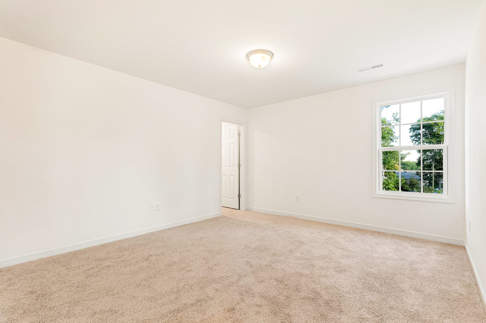 Empty, carpeted room in Poplar Village, Lot 29, with white walls, a window with greenery views, and a ceiling light.
