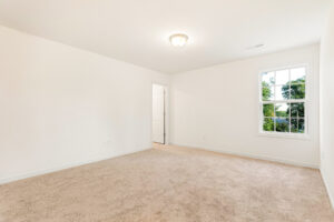 Empty, carpeted room in Poplar Village, Lot 29, with white walls, a window with greenery views, and a ceiling light.