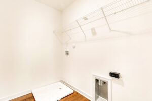 Empty laundry room with white wall, wire shelf, hookups, and wood flooring in Poplar Village Lot 29, ready for appliances.