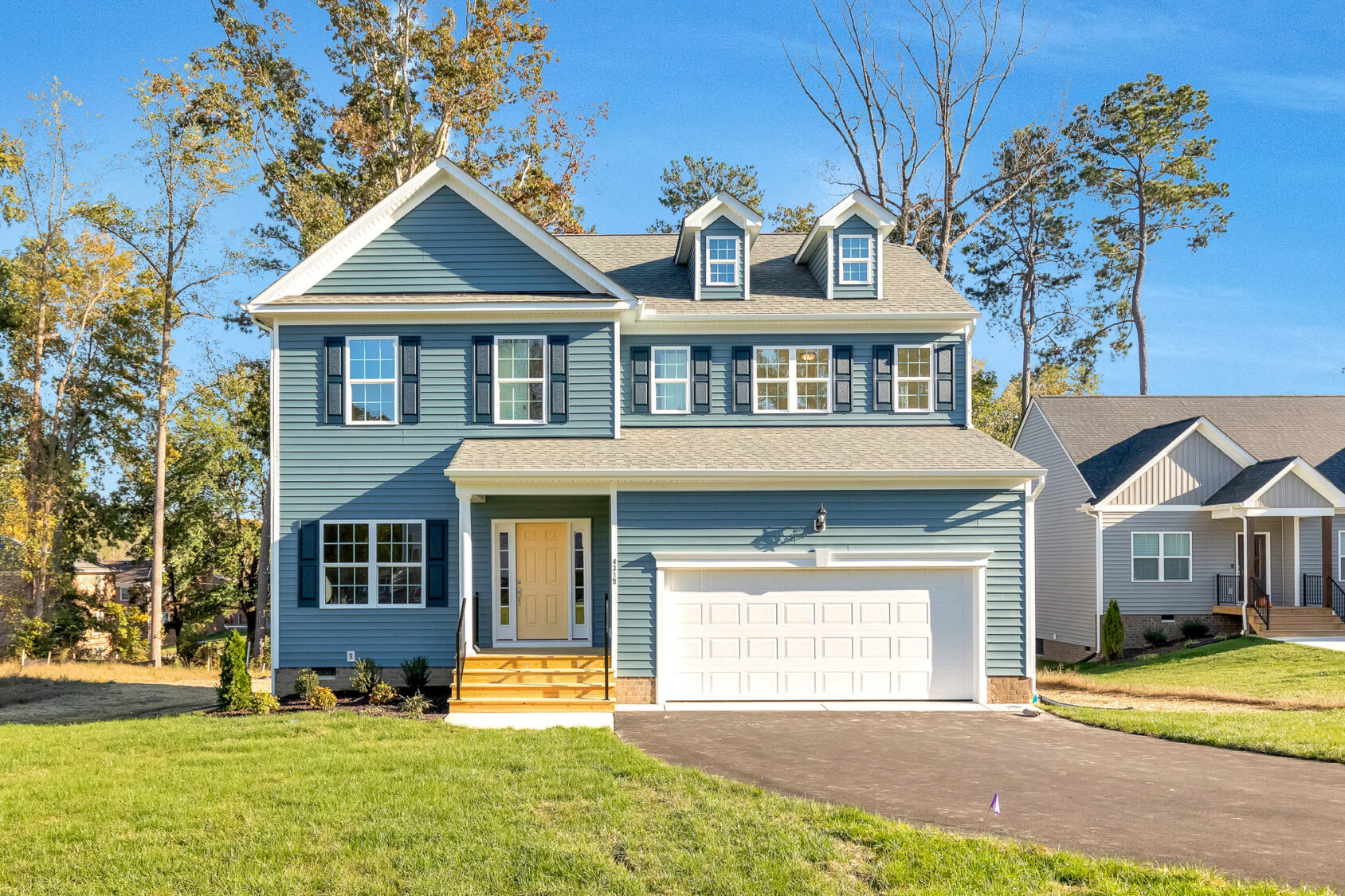 Two-story blue house with white trim on Lot 29 in Poplar Village, featuring a porch and double garage on a sunny street.