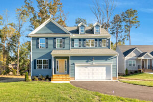 Two-story blue house with white trim on Lot 29 in Poplar Village, featuring a porch and double garage on a sunny street.