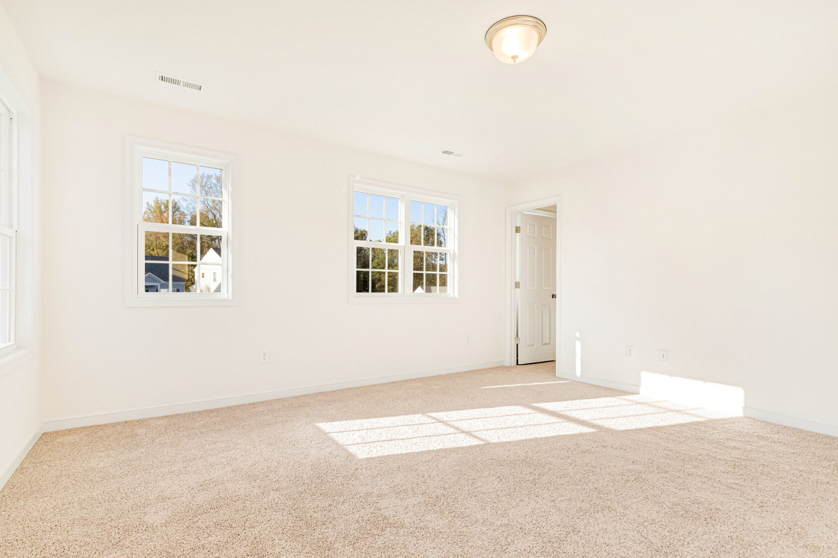 Bright, empty carpeted room in Poplar Village with white walls, three windows, and a ceiling light fixture.