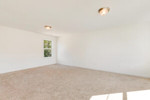 Bright, empty room in Poplar Village’s Lot 23 with beige carpet, white walls, ceiling lights, and sunlit window.