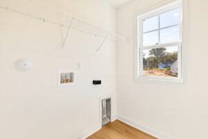 Bright laundry room with white walls, wire shelf, and hookups in Poplar Village Lot 23, window overlooks construction site.
