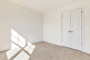 Empty room in Poplar Village, with beige carpet, white walls, double doors, and sunlight casting window shadows.