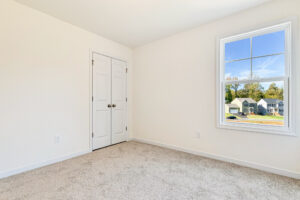 Empty room with beige carpet, white walls, and a closet—located in Poplar Village’s Lot 23 with a window view of houses.