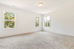 Bright empty room with beige carpet, white walls, and three windows in Poplar Village’s Lot 23 welcoming natural light.