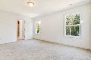 Bright, empty carpeted room in Poplar Village's Lot 23 with two windows, white walls, and doorway to bathroom.