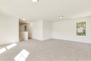 Spacious, empty living room in Poplar Village Lot 23, with beige carpet, white walls, and sunlight through windows.