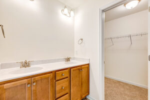 Double sink vanity with wooden cabinets in Poplar Village, beside a walk-in closet with wire shelving and carpeted floor.