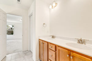 Bathroom with double sinks, wooden cabinets, and a view into the carpeted Poplar Village Lot 23 bedroom through an open door.