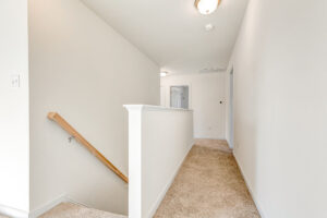 Bright hallway in Poplar Village with beige carpet, white walls, and a wooden stair railing leading to a lower level.