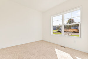 Bright empty room with beige carpet and large window overlooking Poplar Village and Lot 23 on a suburban street.