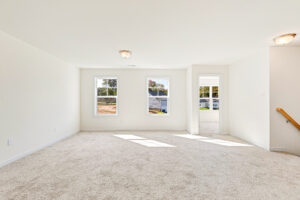 Bright, empty carpeted room in Poplar Village, Lot 23, with white walls and large windows streaming natural light.