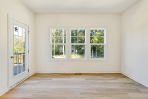 Bright empty room with large windows and light wood flooring in Poplar Village’s Lot 23, overlooking trees outside.