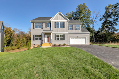 Two-story gray house on Lot 23 in Poplar Village with a front porch, attached garage, and sloped driveway on a sunny day.