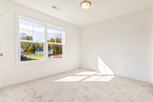 Bright, empty room with beige carpet and large window in Poplar Village’s Lot 23; houses visible outside.