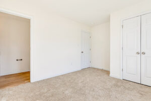Empty beige carpeted room in Poplar Village with white walls, two doors, and an open doorway to Lot 23’s wood floor.
