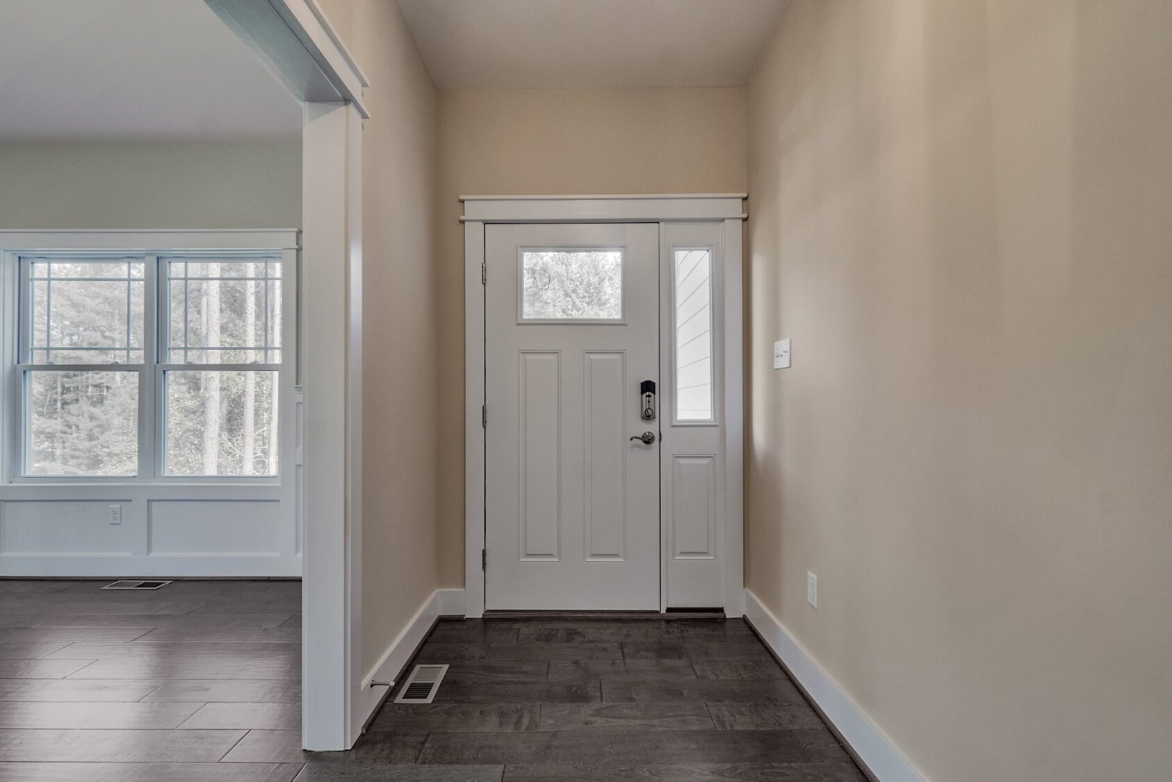 A white front door with glass panels in a beige entryway at Poplar Village, beside a room with large windows and dark flooring.