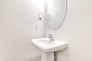 A white pedestal sink with a round mirror in the minimalist Poplar Village Lot 13 bathroom.