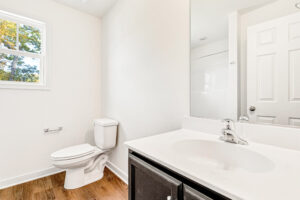 Bright Poplar Village bathroom with a white toilet, sink, large mirror, wood floors, and sunlight from a window.