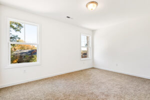 Bright, empty room in Poplar Village with beige carpet, white walls, two windows, and a ceiling light fixture.
