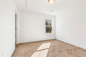 Bright, empty room with beige carpet in Poplar Village, Lot 13—features white walls, window, door, and ceiling light.