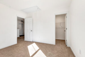 Bright, empty bedroom in Poplar Village’s Lot 13 with beige carpet, white walls, and open doors to closet and hallway.