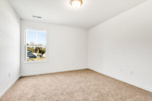 Empty room in Poplar Village with beige carpet, white walls, ceiling light, and window overlooking houses.