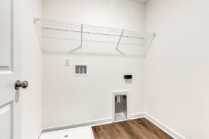 Empty laundry room in Poplar Village’s Lot 13 with white walls, wire shelf, hookups, and brown wood floor.