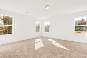 Bright, empty room in Poplar Village with beige carpet, white walls, and three large windows letting in sunlight.