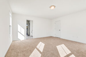 Bright, empty bedroom in Poplar Village’s Lot 13 with beige carpet, white walls, and sunlight streaming through two windows.