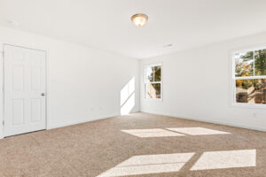 Empty, bright room in Poplar Village with beige carpet, white walls, two windows, and a closed white door.
