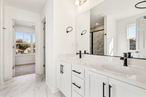 Modern white bathroom in Poplar Village with double sinks, marble countertop, and black fixtures; bedroom visible beyond.