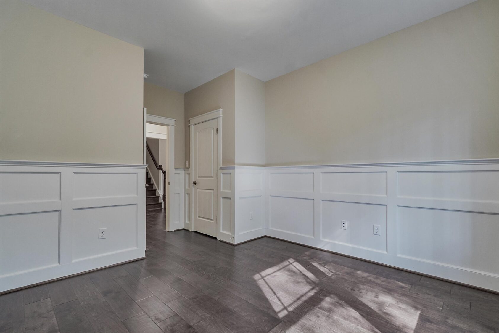 Empty room in Poplar Village with beige walls, white wainscoting, dark wood floors, and a doorway to the hallway.