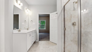 Modern Somerset Hill bathroom with double sinks, large mirror, glass shower, and view into a Bethany bedroom.