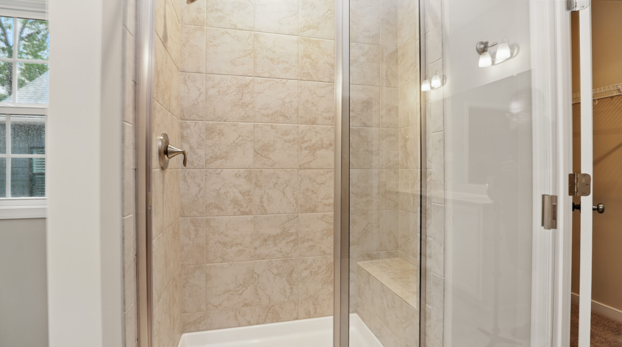 Glass-enclosed shower with beige tiled walls, built-in bench, and silver fixtures in Bethany’s Somerset Hill home.